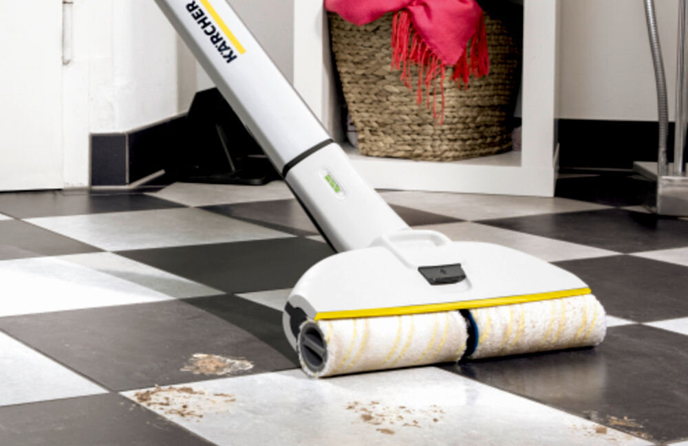 Na obrazie widoczny jest elektryczny mop z logo KARCHER, czyszczący czarno-białą podłogę z zabrudzeniami. W tle znajduje się kosz wiklinowy z różowym materiałem.