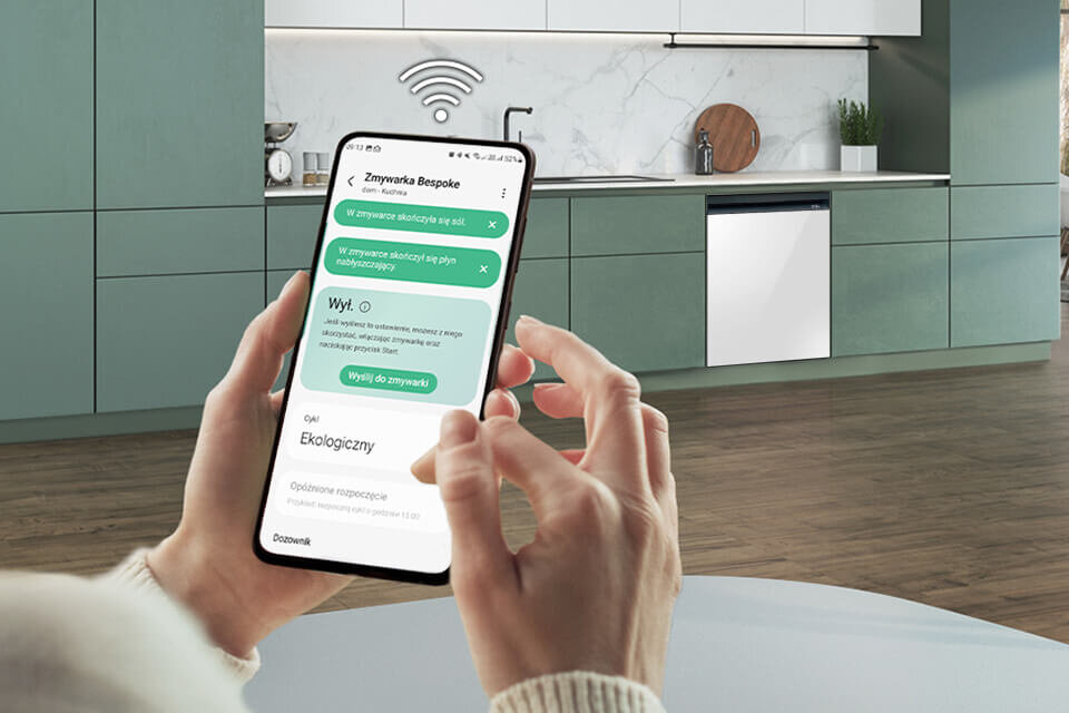 Osoba trzyma smartfon z aplikacją \'Zmywarka Bespoke\', na ekranie widoczne są opcje sterowania zmywarką. W tle znajduje się kuchnia z zabudowaną zmywarką i symbolem Wi-Fi nad nią.
