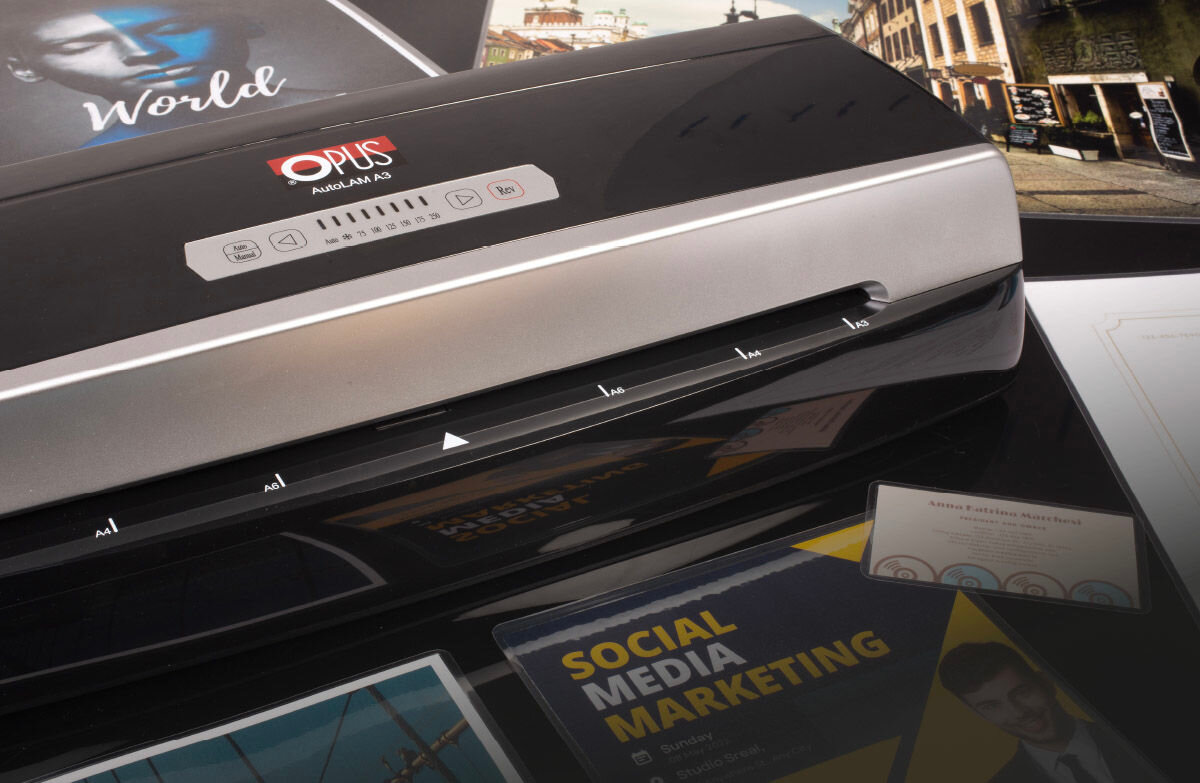 Na obrazie widoczny jest laminator z logo \'OPUS\' i napisem \'AutoLAM A3\'. Wokół urządzenia znajdują się różne materiały drukowane, w tym broszura z napisem \'SOCIAL MEDIA MARKETING\'.
