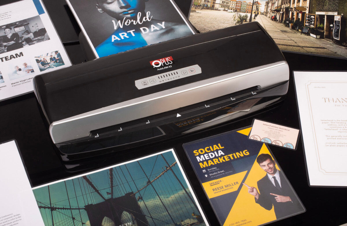 Na obrazie znajduje się laminator z logo \'OPUS\' oraz różne dokumenty, w tym okładka z napisem \'World ART DAY\' i książka \'SOCIAL MEDIA MARKETING\'.