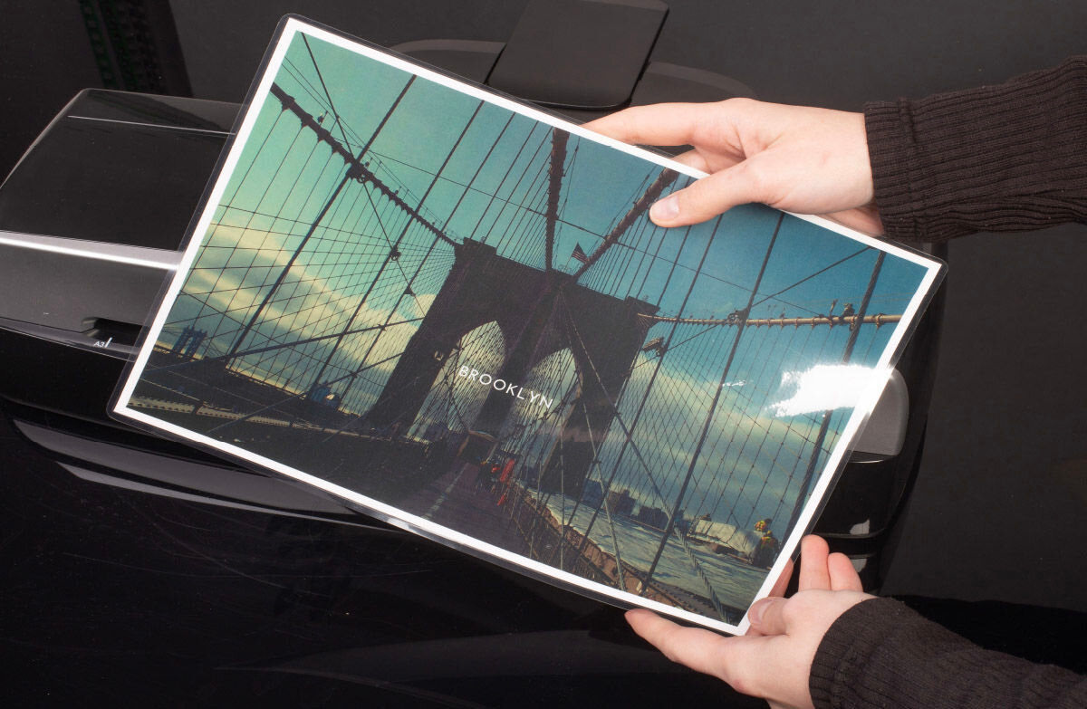 Osoba trzyma zalaminowaną fotografię mostu z napisem \'BROOKLYN\', a w tle widoczny jest laminator.