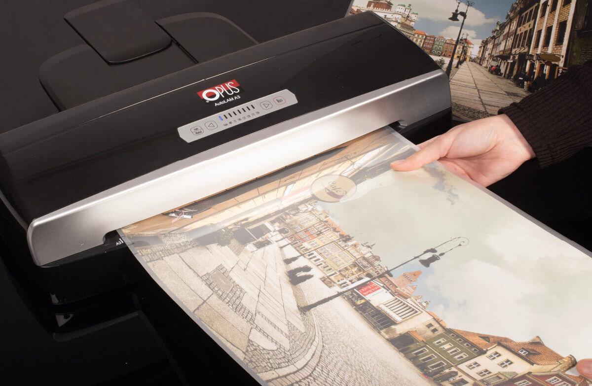 Laminator z logo \'OPUS autoLAM A3\' laminuje kolorowy obrazek przedstawiający ulicę z budynkami. Osoba trzymająca obrazek wkłada go do urządzenia.