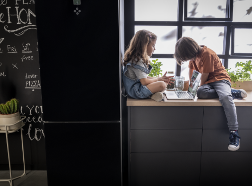 Dwoje dzieci siedzi na blacie kuchennym obok czarnej lodówki, przeglądając coś na laptopie. W tle widać okno i tablicę z napisami, a na blacie stoją doniczki z roślinami.