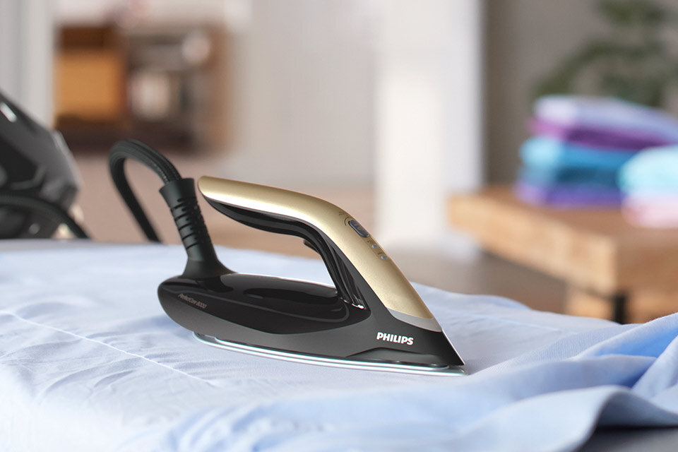 Na obrazie widoczny jest generator pary z napisem \'PHILIPS\' na stopie, umieszczony na niebieskim materiale. W tle znajdują się złożone kolorowe tkaniny na stole.