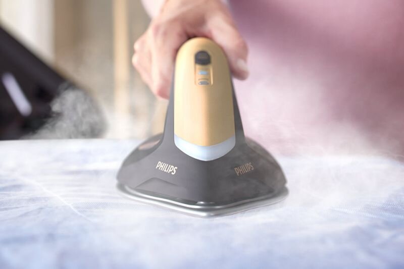 Osoba prasuje tkaninę za pomocą generatora pary z widocznym logo PHILIPS. Urządzenie emituje parę podczas prasowania.