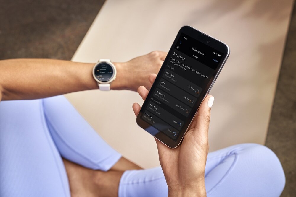 Osoba siedzi na macie do ćwiczeń, trzymając w ręku smartfon z aplikacją wyświetlającą dane zdrowotne. Na nadgarstku ma założony sportowy zegarek z okrągłą tarczą.