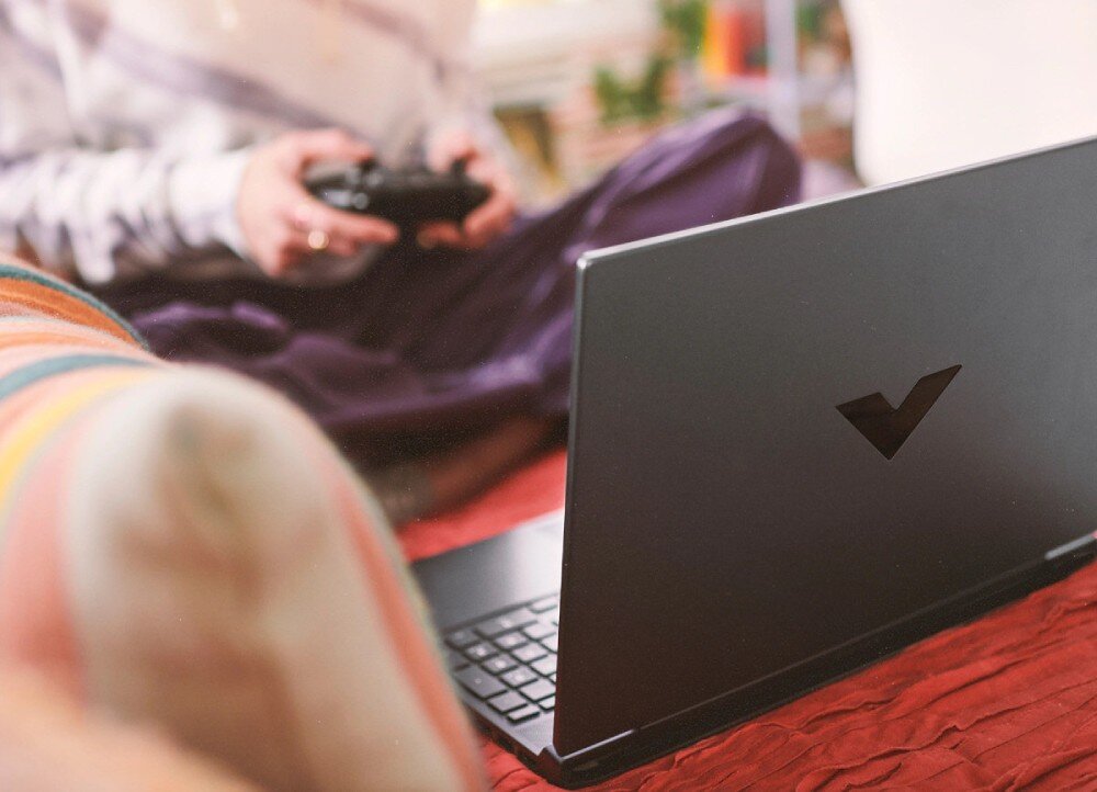 Osoba siedzi na łóżku trzymając kontroler do gier, przed nią znajduje się otwarty laptop z widocznym logo w kształcie litery V na obudowie.