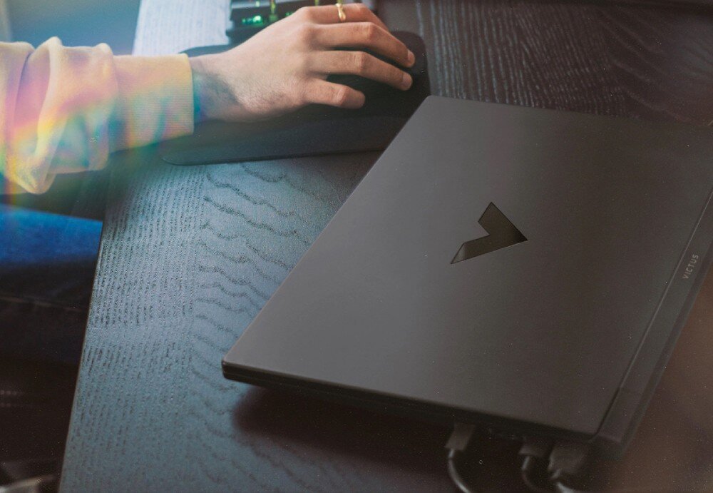 Na ciemnym biurku leży zamknięty laptop z logo w kształcie litery \'V\'. Obok znajduje się ręka na myszce komputerowej.