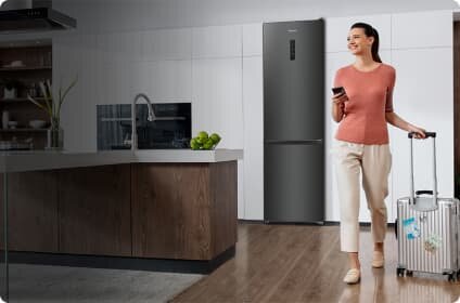 Kobieta trzymająca telefon i walizkę stoi w nowoczesnej kuchni z czarną lodówką HISENSE. Na blacie kuchennym znajdują się zielone jabłka.