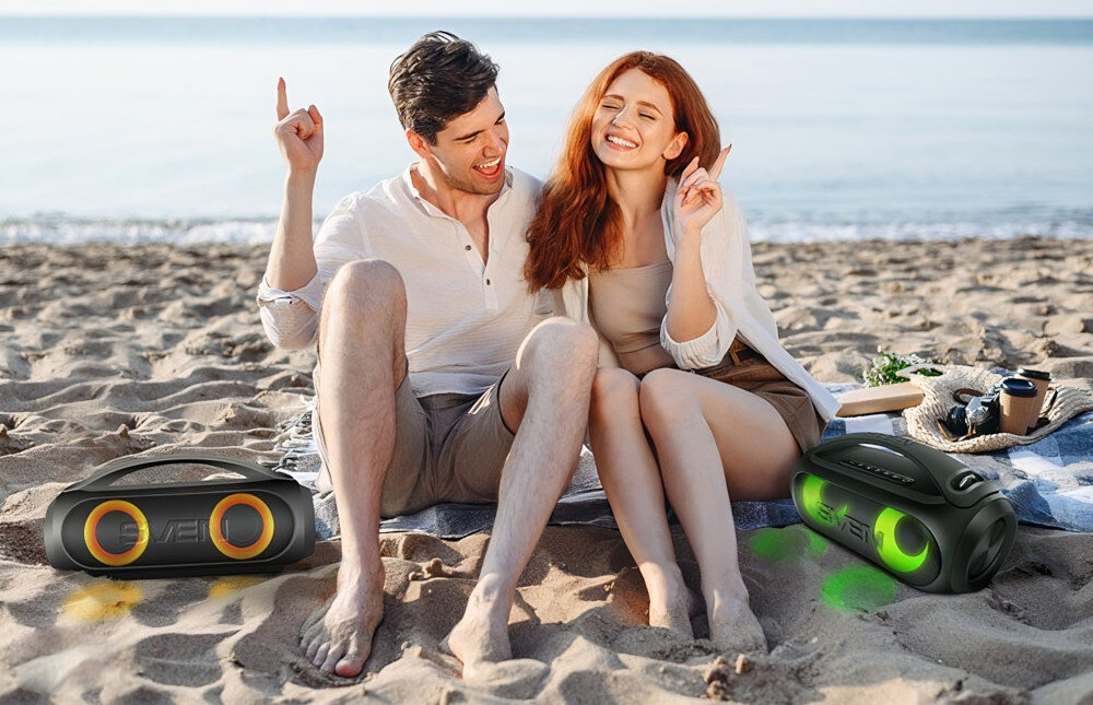 Na plaży siedzi para, obok której znajdują się dwa czarne głośniki mobilne z kolorowymi elementami. W tle widać morze i jasne niebo.