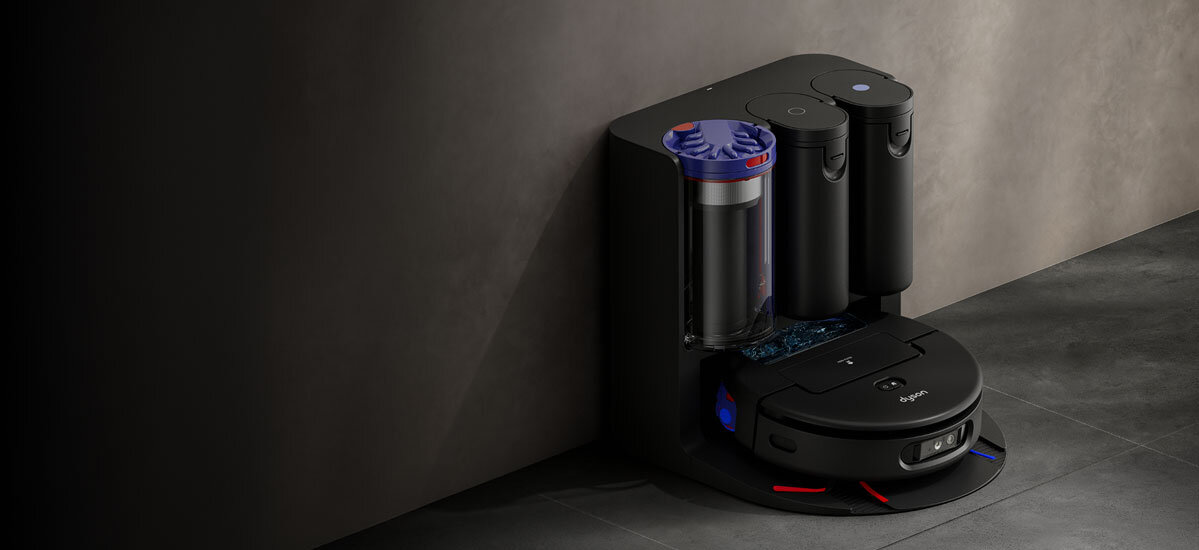 Na obrazie widoczny jest robot sprzątający w stacji dokującej z dwoma cylindrycznymi pojemnikami. Na robocie znajduje się napis \'Dyson\'.