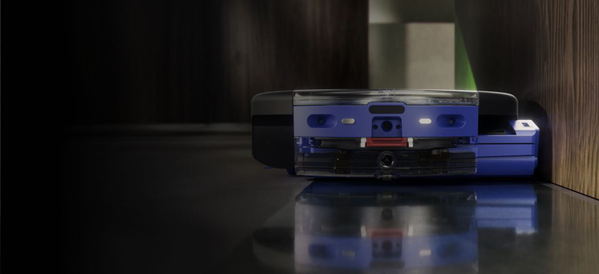 Robot sprzątający w kolorze niebieskim znajduje się przy ścianie na ciemnej powierzchni. Widać jego przednią część z czujnikami i elementami konstrukcyjnymi.