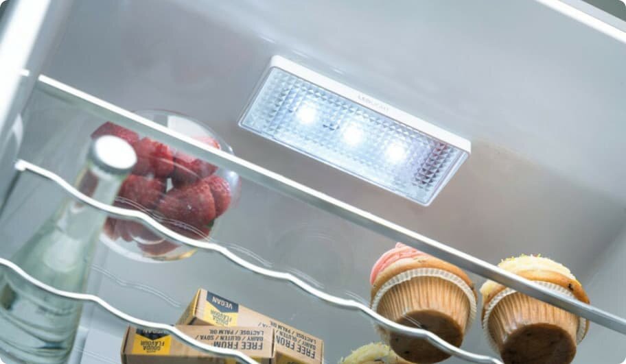 Wnętrze lodówki z oświetleniem LED, widoczne są półki z metalowymi prętami. Na półkach znajdują się butelka, pojemnik z malinami i dwa muffiny.