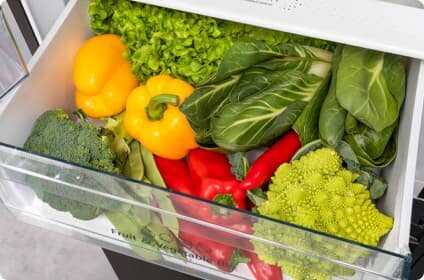 Szuflada lodówki wypełniona różnymi warzywami, w tym papryką, brokułem, sałatą i romanesco. Na szufladzie widoczny jest napis \'Fruit & Veg Zone\'.