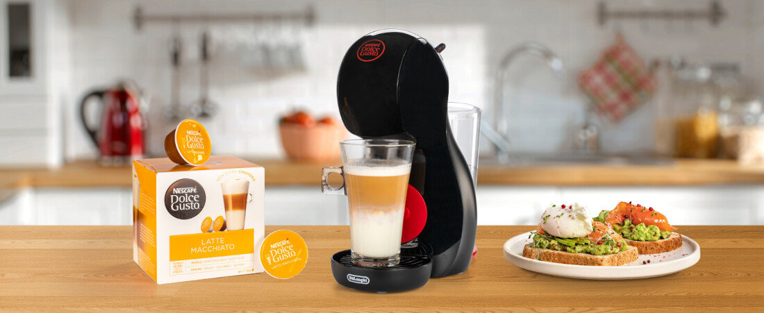 Na blacie kuchennym stoi czarny ekspres do kawy z logo \'Dolce Gusto\', obok znajduje się szklanka z kawą latte macchiato. Po lewej stronie widoczna jest paczka kapsułek \'Latte Macchiato\' marki \'Nescafe Dolce Gusto\'.