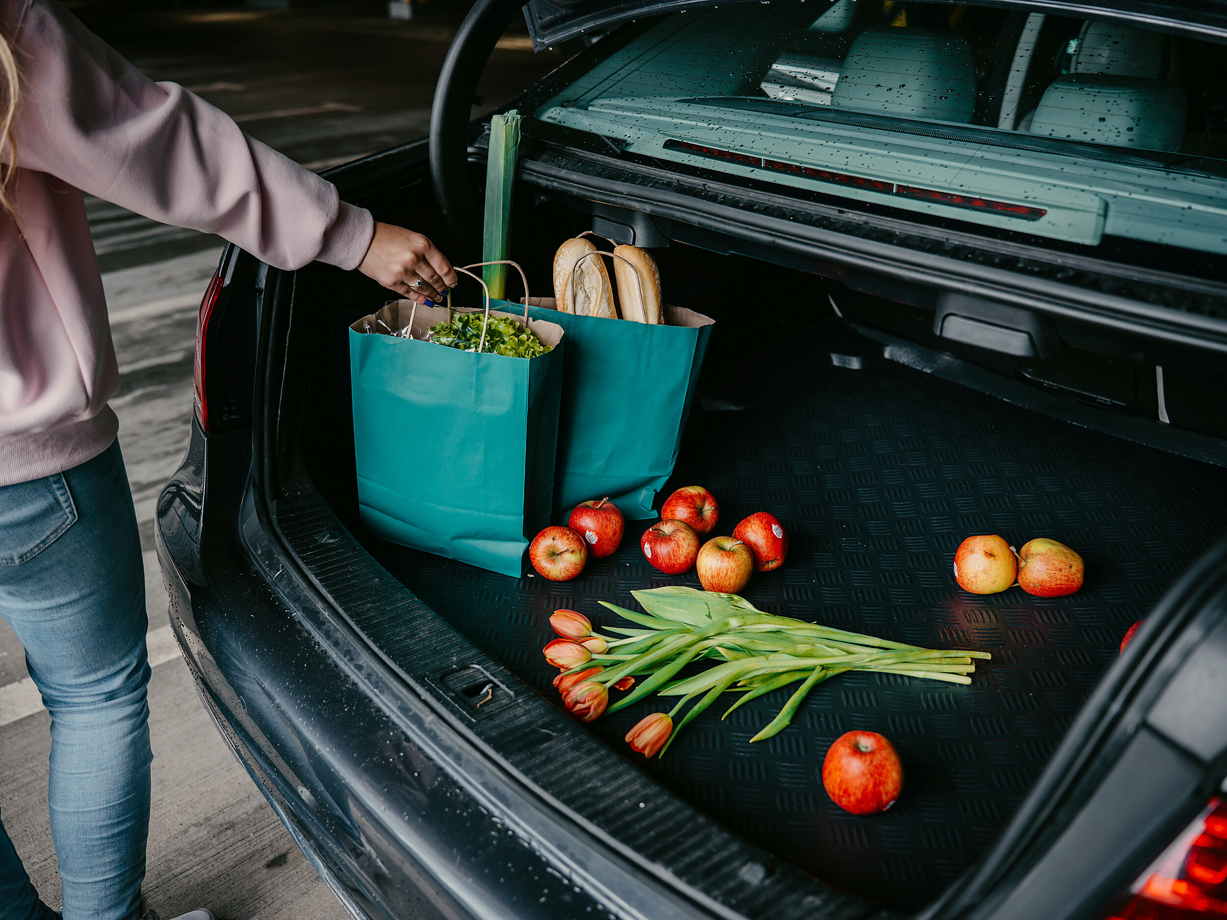 Otwarty bagażnik samochodu z matą, na której leżą jabłka i tulipany. Obok znajdują się torby z zakupami, a osoba w różowej bluzie sięga po jedną z nich.
