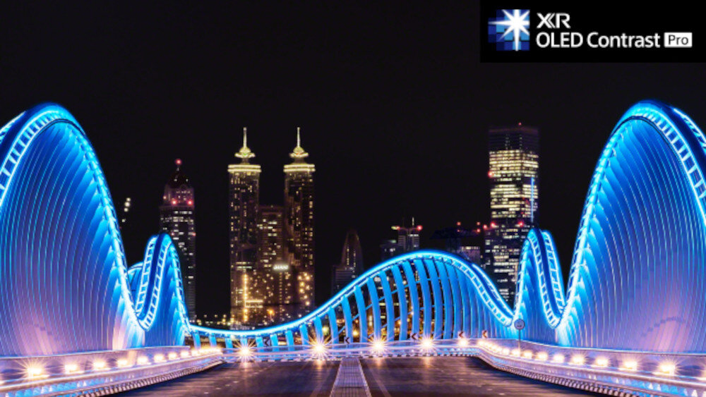 Nocna panorama miasta z futurystycznym mostem oświetlonym niebieskimi światłami. W prawym górnym rogu widoczne jest logo z napisem \'XR OLED Contrast Pro\'.