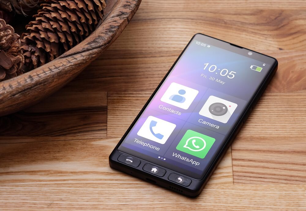 Smartfon leży na drewnianym stole obok miski z szyszkami. Na ekranie widoczne są ikony aplikacji: Contacts, Camera, Telephone, WhatsApp oraz godzina 10:05 i data Fri, 30 May.