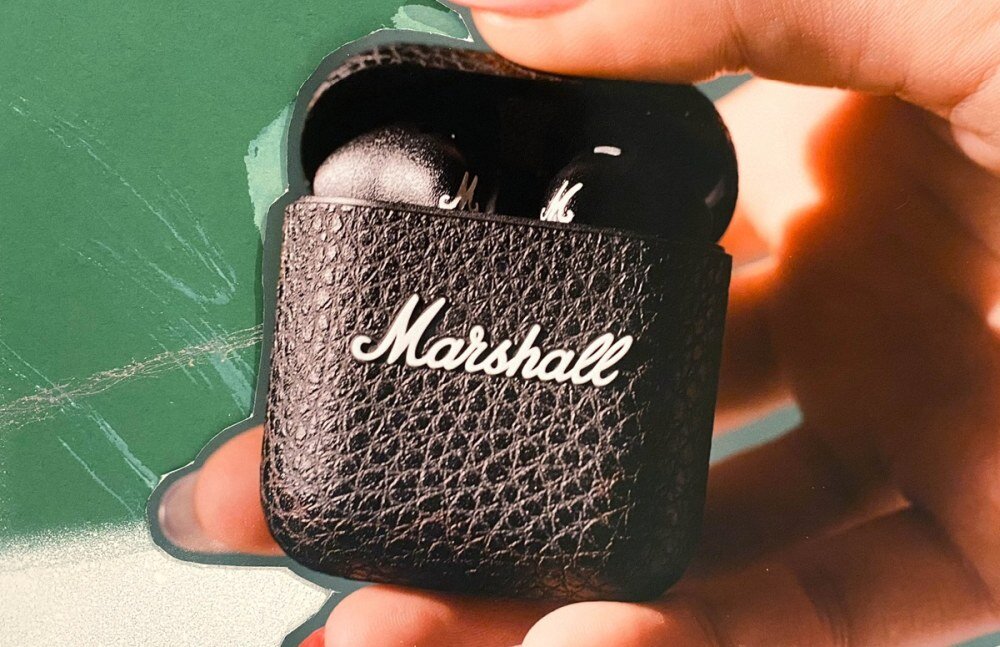 Czarna, teksturowana etui na słuchawki z widocznym logo \'Marshall\' trzymana w dłoni. Wewnątrz znajdują się czarne słuchawki douszne.