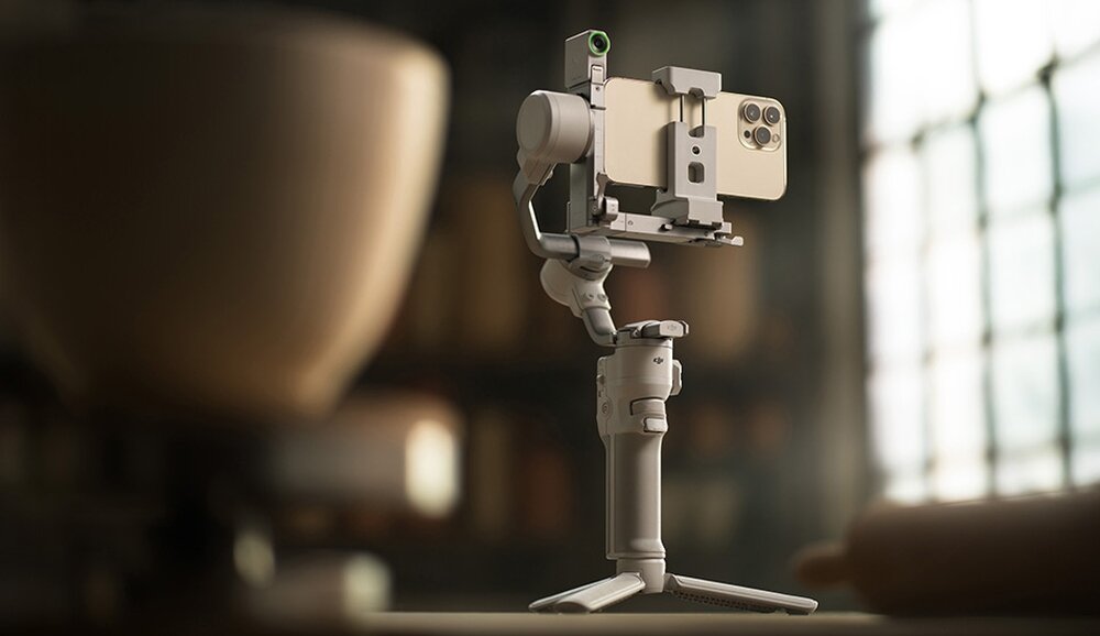 Na obrazie widoczny jest szary gimbal z zamocowanym smartfonem, ustawiony na statywie. W tle znajduje się rozmyte wnętrze z dużym oknem.