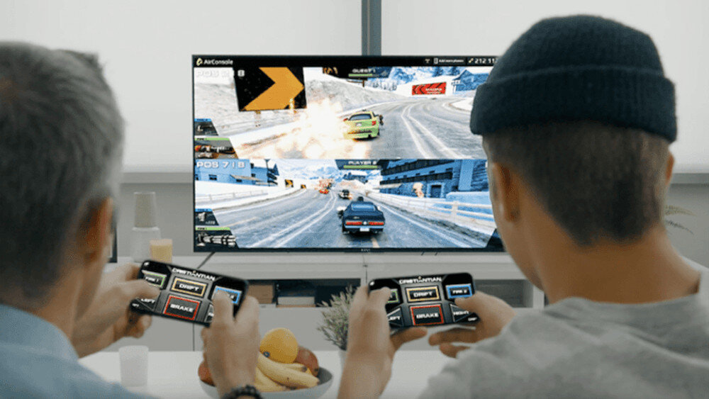Dwóch mężczyzn gra w grę wyścigową na telewizorze, używając smartfonów jako kontrolerów. Na ekranie widać zimowy krajobraz i samochody na torze wyścigowym.