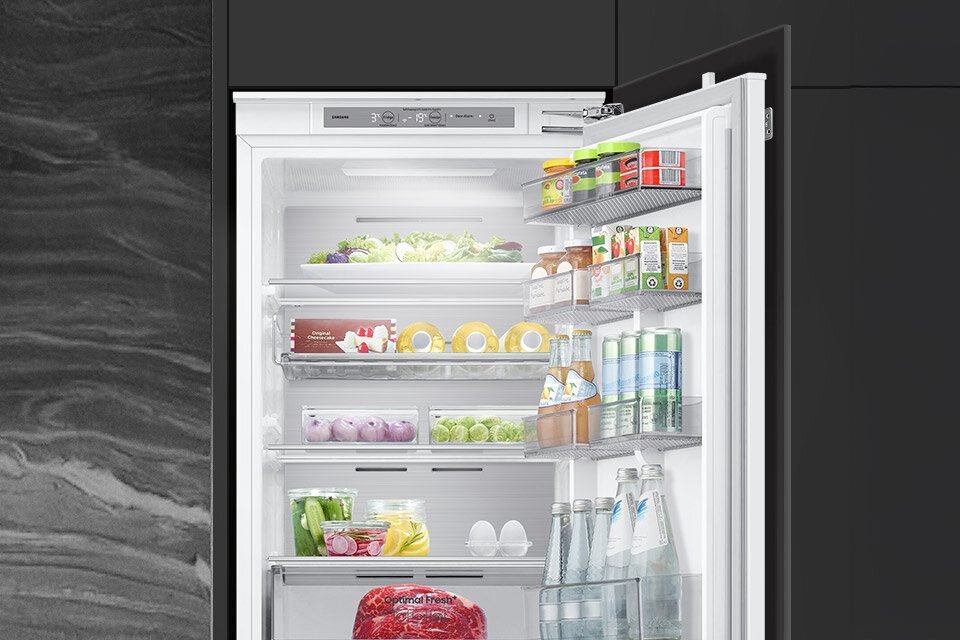 Otwarta lodówka z widocznymi półkami wypełnionymi różnorodnymi produktami spożywczymi, takimi jak warzywa, napoje i jajka. Na drzwiach lodówki znajdują się dodatkowe półki z napojami i kartonami.