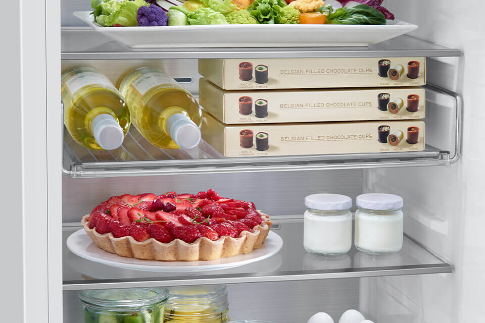 Wnętrze lodówki z półkami, na których znajdują się różne produkty spożywcze, w tym butelki z napojami, pudełka z czekoladkami, tarta z truskawkami oraz słoiki z jogurtem. Na górnej półce widoczny jest talerz z warzywami, a na dolnej jajka i słoiki z przetworami.