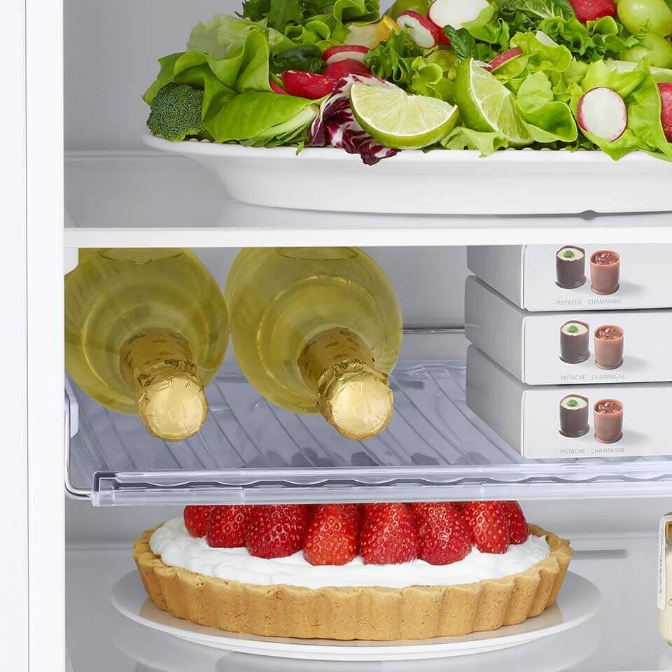 W lodówce znajdują się dwie butelki wina, ciasto z truskawkami oraz pudełka z czekoladkami. Na górnej półce widoczna jest sałatka z warzywami i plasterkami limonki.