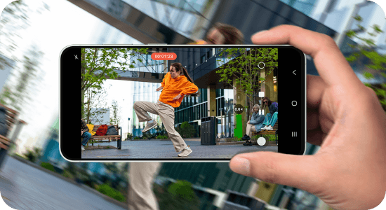 Osoba trzyma smartfon, na ekranie którego widać nagranie wideo przedstawiające osobę w pomarańczowej bluzie wykonującą taneczny ruch na tle miejskiego krajobrazu. Na ekranie widoczny jest licznik czasu nagrania \'00:01:23\'.