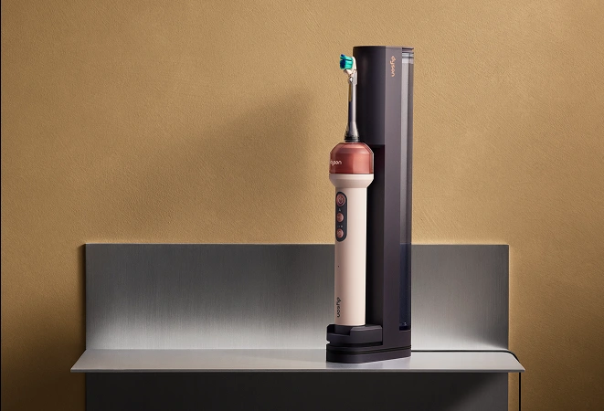 Яскраво-рожева електрична зубна щітка Dyson CameraJet стоїть у фіолетовій док-станції на сірій стільниці.