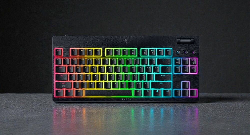Klawiatura z podświetleniem RGB, z widocznym logo producenta na górze. Klawisze są ułożone w standardowym układzie, a obudowa jest czarna.