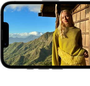 Smartfon wyświetla zdjęcie osoby stojącej na tle górskiego krajobrazu. Osoba ubrana jest w żółty sweter, a w tle widać drewnianą konstrukcję i niebo z chmurami.