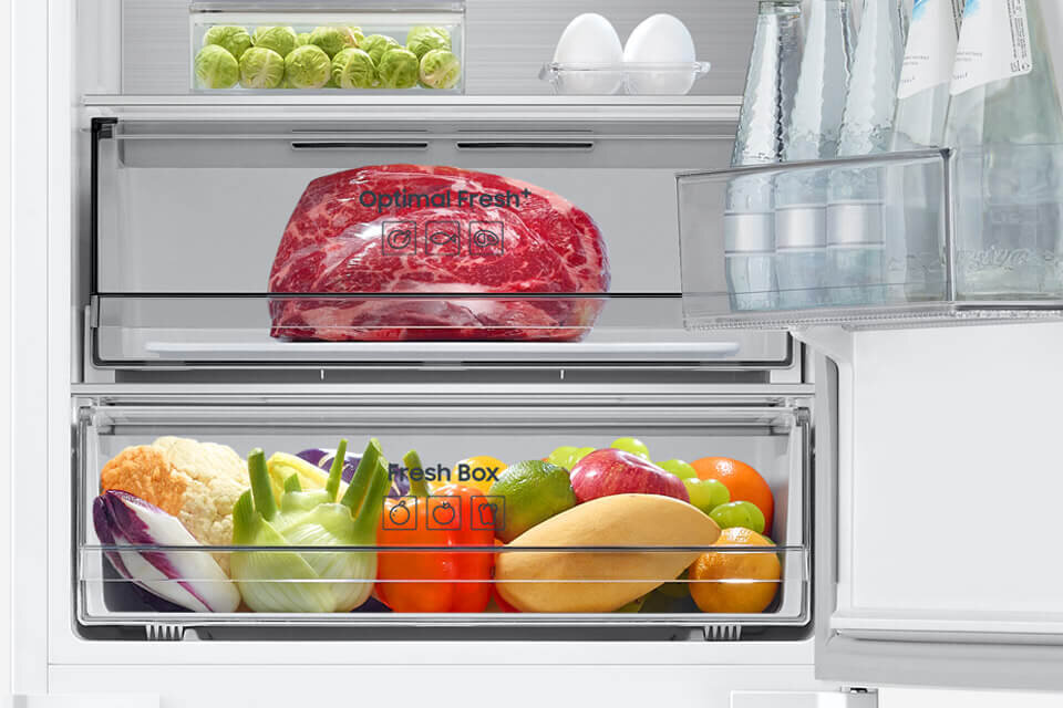 Wnętrze lodówki z widocznymi szufladami oznaczonymi jako \'Optimal Fresh+\' i \'Fresh Box\', wypełnionymi mięsem oraz różnorodnymi warzywami i owocami. Na drzwiach znajdują się butelki z wodą, a na półce jajka i brukselka.