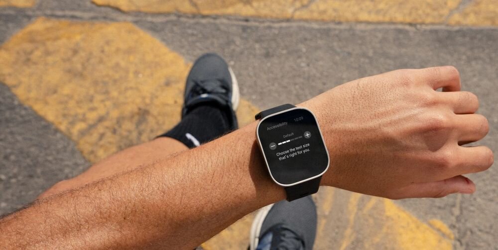 Osoba siedzi na zewnątrz, nosząc smartwatch na nadgarstku. Na ekranie zegarka widoczny jest tekst: \'Choose the best size that is right for you\'.