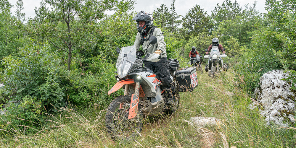 Trzech motocyklistów jedzie po wąskiej, trawiastej ścieżce otoczonej drzewami i krzewami. Motocykle są wyposażone w kufry i osłony, a motocykliści mają na sobie kaski i odzież ochronną.