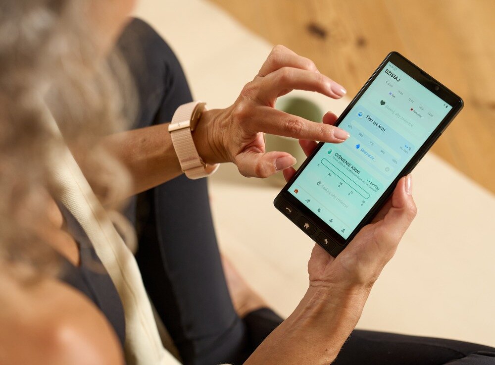 Osoba trzyma smartfon, na ekranie widoczny jest interfejs aplikacji z różnymi opcjami i ustawieniami. Osoba używa palca do nawigacji po ekranie dotykowym.