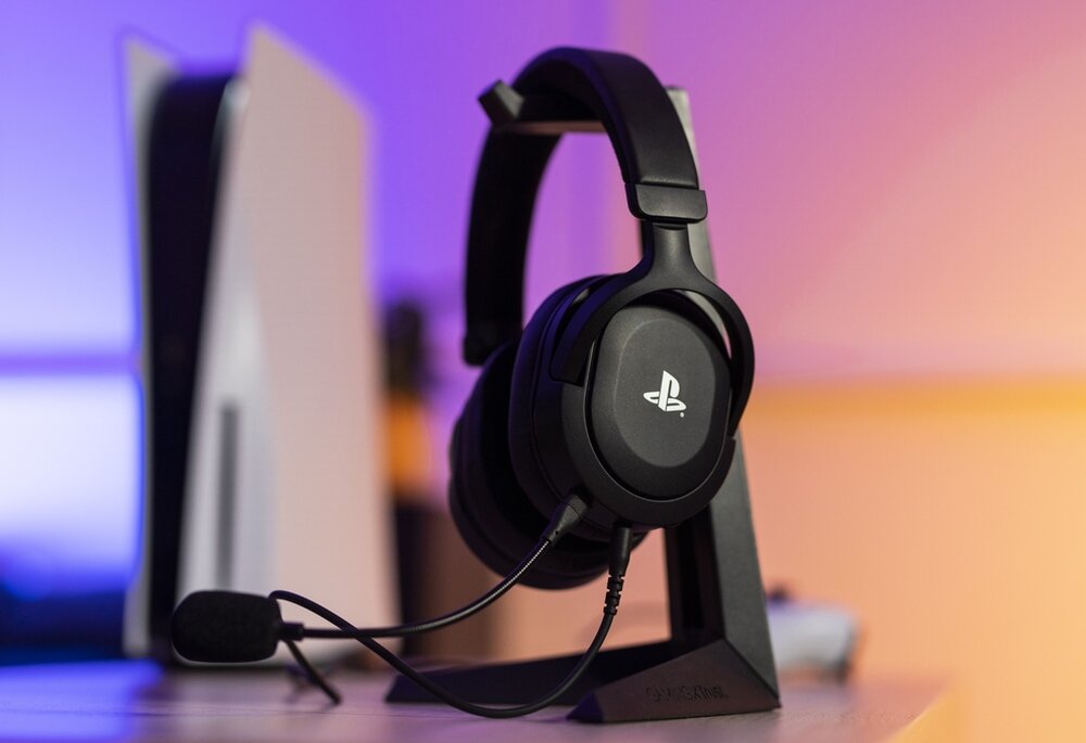 Czarne słuchawki nauszne z mikrofonem stoją na stojaku, obok konsoli do gier. Na słuchawkach widoczne jest logo PlayStation.