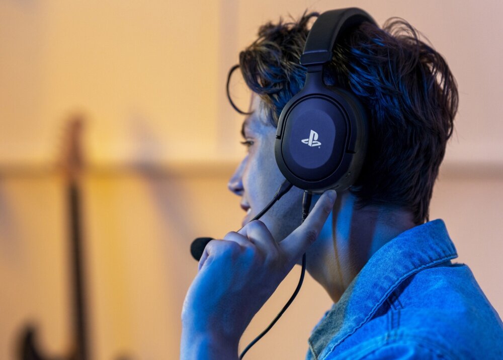 Osoba nosi czarne słuchawki z widocznym logo PlayStation. W tle rozmyty obraz gitary.