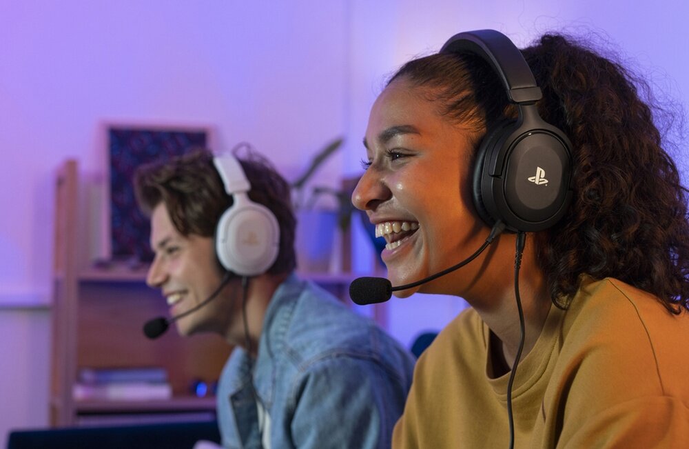 Dwie osoby noszące słuchawki z mikrofonem, jedna z nich ma czarne słuchawki z logo PlayStation. W tle widać półkę z książkami i roślinami.