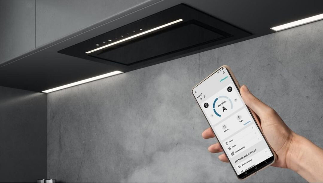 Na obrazie widoczny jest nowoczesny okap kuchenny zamontowany w szafce, z panelem dotykowym i oświetleniem LED. Osoba trzyma smartfon z aplikacją do sterowania urządzeniem.