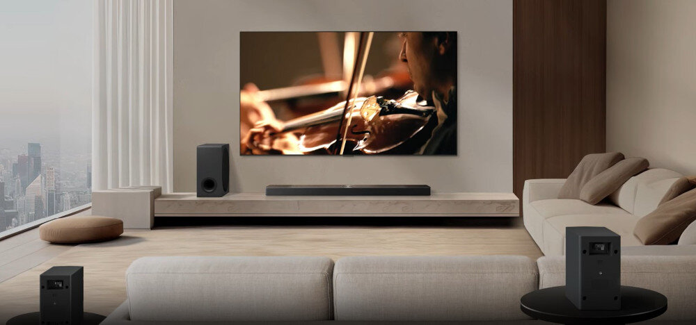 W nowoczesnym salonie znajduje się duży telewizor wyświetlający obraz skrzypiec, a pod nim umieszczony jest soundbar. W pomieszczeniu widoczne są również głośniki i wygodna sofa.