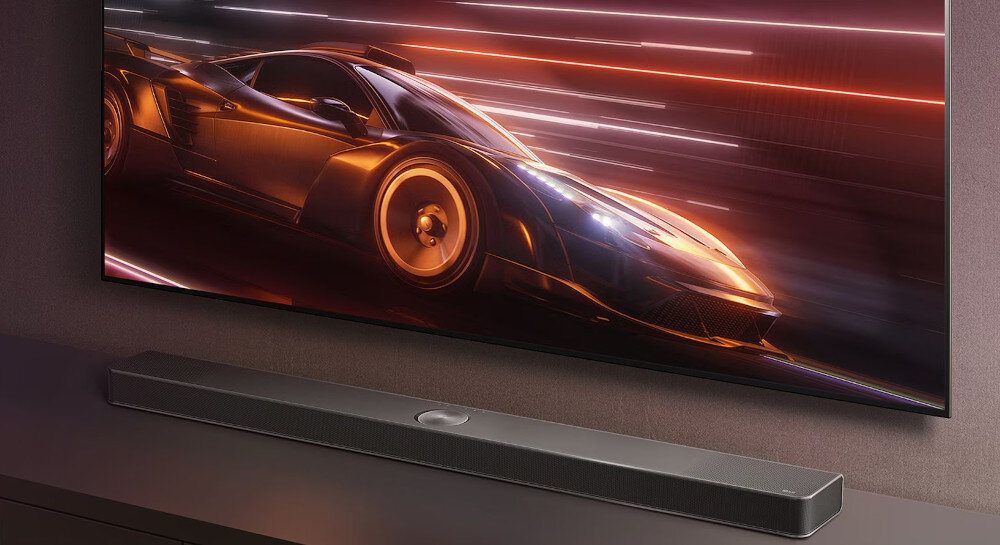 Na obrazie widoczny jest czarny soundbar umieszczony pod dużym telewizorem, na którym wyświetlany jest dynamiczny obraz samochodu wyścigowego. Soundbar ma prosty, nowoczesny design.