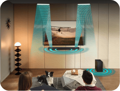 Pokój z telewizorem na ścianie, przed którym siedzą dwie osoby i pies. Obok telewizora znajduje się soundbar i subwoofer, a z głośników emitowane są wizualizacje dźwięku.