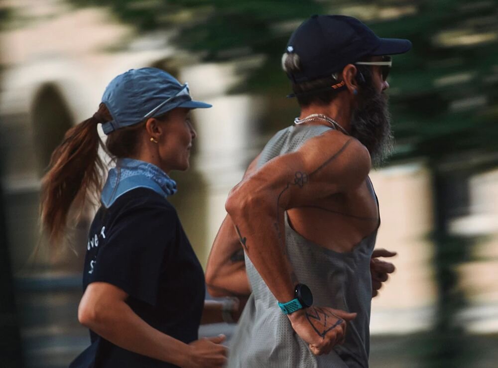 Dwie osoby biegną obok siebie, jedna z nich ma na nadgarstku zegarek sportowy. Obie osoby są ubrane w sportowe stroje i czapki.