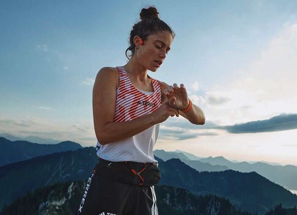 Osoba w sportowym stroju stoi na tle górskiego krajobrazu, patrząc na swoje nadgarstkowe urządzenie. Na uszach ma czarne słuchawki.