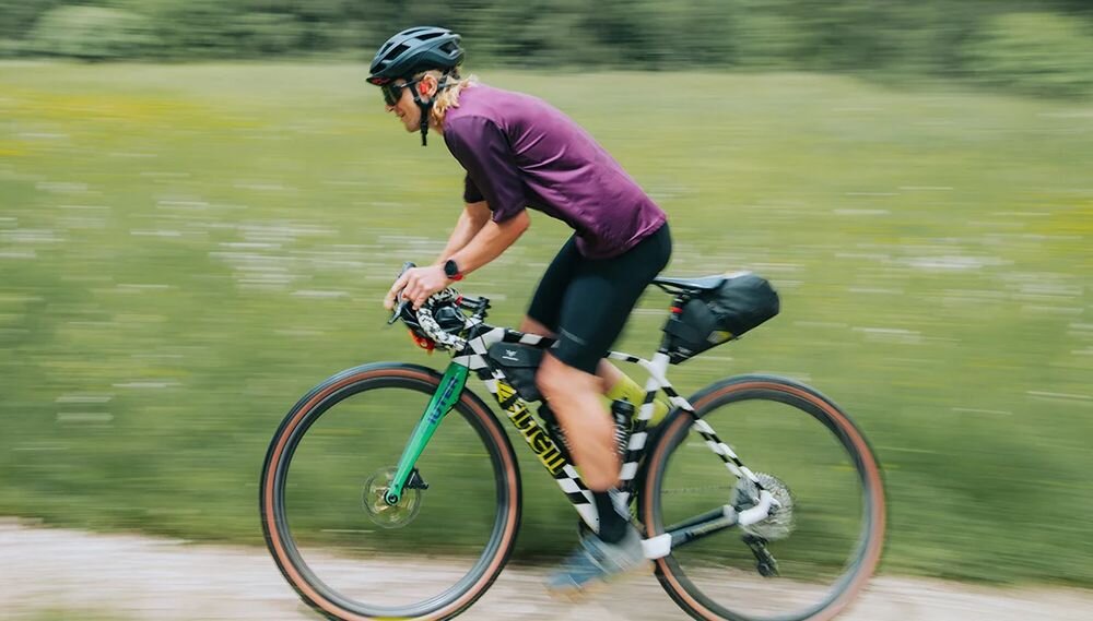 Osoba na rowerze jedzie po ścieżce w otoczeniu zieleni, ubrana w kask i sportowy strój. Rower ma kolorowe elementy i torbę zamocowaną z tyłu.