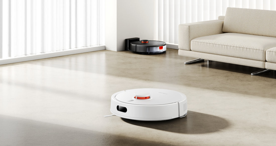 Na podłodze w jasnym pomieszczeniu znajduje się biały robot sprzątający. W tle widać ciemny robot przy stacji dokującej oraz jasną sofę.