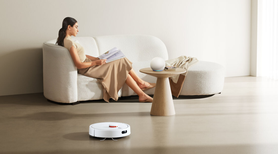 Na obrazie widać biały robot sprzątający na podłodze w nowoczesnym wnętrzu. W tle znajduje się osoba siedząca na jasnej sofie, trzymająca książkę, obok stoi drewniany stolik z kulistą lampą.