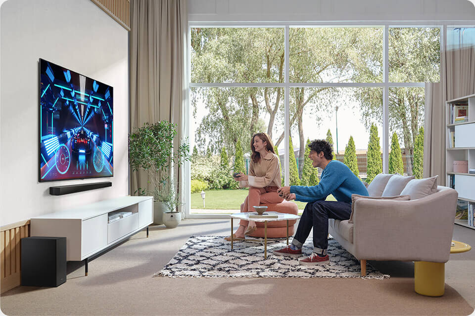 960c640- hw-b750f sound quality game mode 2 W salonie znajduje się telewizor z wyświetlanym kolorowym obrazem oraz soundbar umieszczony pod nim. Na kanapie siedzą dwie osoby, a za nimi widać duże okno z widokiem na ogród.