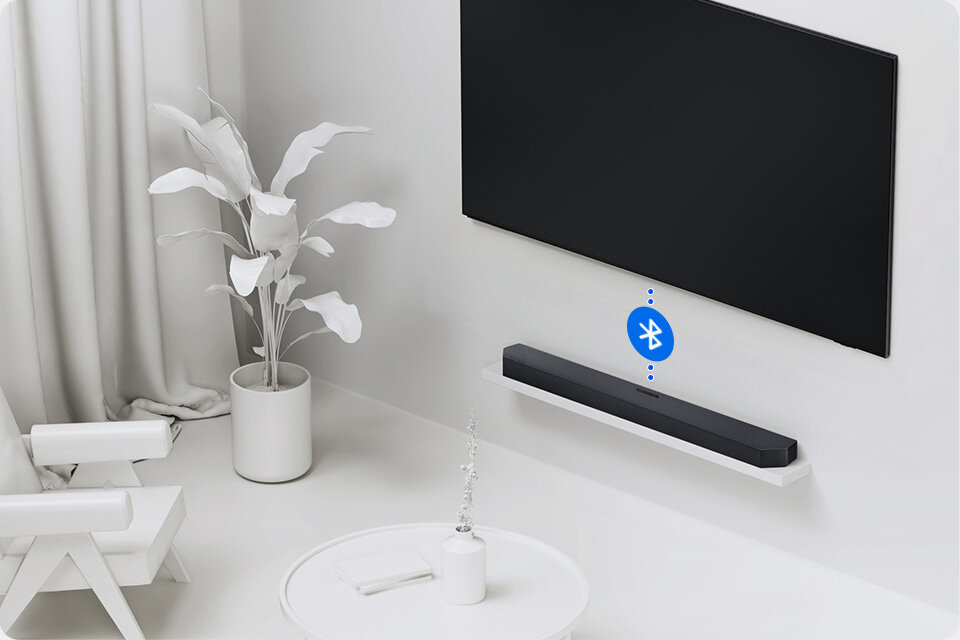 960c640- wireless-tv-connection 2 Na obrazie widać czarny soundbar umieszczony na białej półce pod dużym telewizorem. Obok soundbara znajduje się niebieska ikona Bluetooth, a w tle widoczna jest biała roślina w doniczce i biały fotel.
